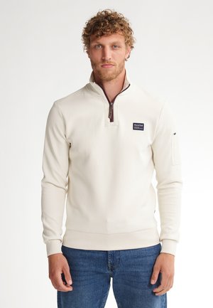 Helle beige Half-Zip-Pullover aus weichem Material, mit einem kastanienbraunen Reißverschluss und Patch-Label auf der Brust, sowie eng anliegenden Bündchen und Saum.