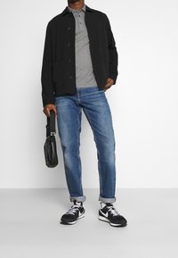 Svart knapplös jacka över en grå polo, blå jeans och svarta sneakers. Accessoarer inkluderar en svart axelväska med medelstruktur.