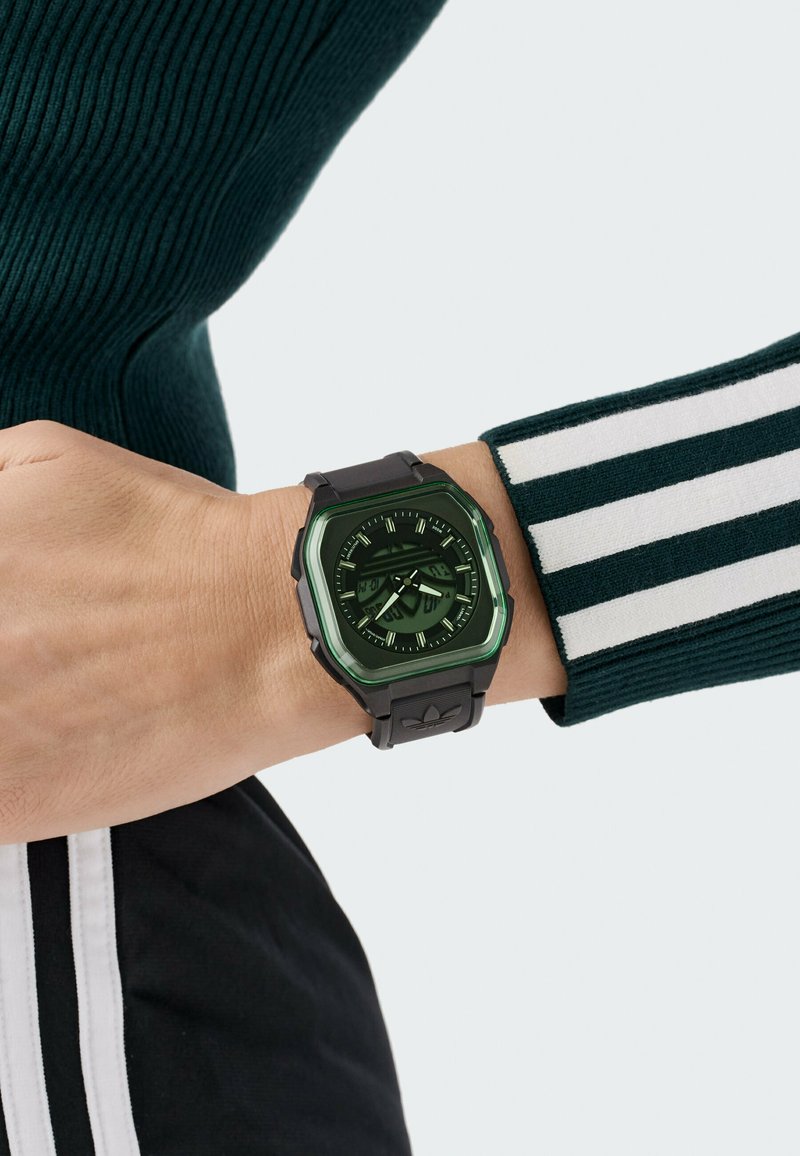 Pols met zwarte digitaal-analoog horloge met groene accenten, gecombineerd met een donkergroene geribbelde trui en zwarte broek met witte strepen.