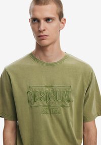 Camiseta de algodón verde con el logotipo bordado "DESIGUAL" y "EST. 1984". Cuello redondo, mangas cortas, corte ligeramente relajado y textura suave.