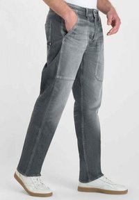 Homme portant un jean gris bicolore avec des poches latérales et des baskets blanches à semelles en caoutchouc, se tenant avec une main dans la poche.