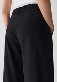 Pantalons noirs avec une texture lisse, une taille élastique, des poches latérales et une poche arrière unique, présentant un design à jambes larges.