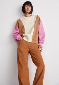 Sweatshirt colorblock en crème, marron et rose, en tissu doux ; associé à un pantalon en velours côtelé à jambe large de couleur rouille.
