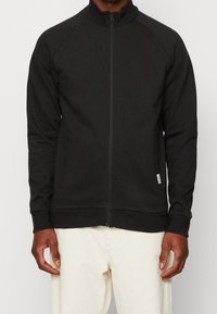 Veste zippée noire avec un col haut, des manches raglan et des poches latérales. Fabriquée dans un tissu lisse et extensible avec des poignets ajustés.