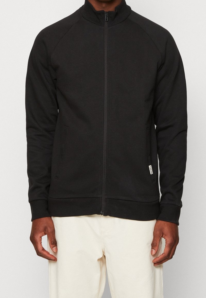 Veste zippée noire avec un col haut, des manches raglan et des poches latérales. Fabriquée dans un tissu lisse et extensible avec des poignets ajustés.