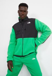 Fleece striukė su juodu viršutiniu dalyku ir ryškiai žaliu apatinio dalimi, su užtrauktukais ir krūtinės kišenėmis, logotipas krūtinėje ir rankovėje.