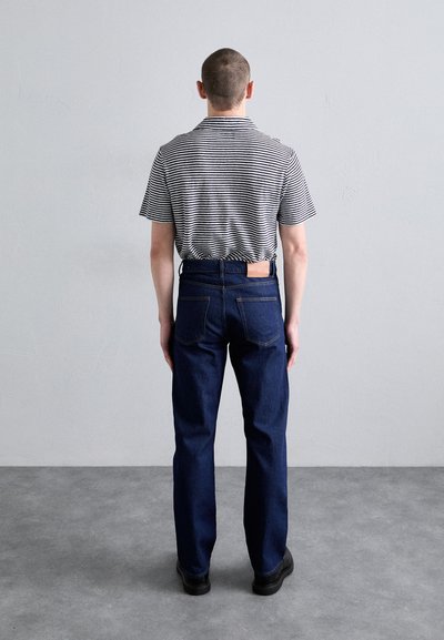 Camisa de manga corta a rayas con cuello, combinada con jeans de mezclilla azul oscuro. Bolsillos traseros visibles y una etiqueta en la cinturilla.