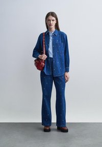 Femme portant une veste et un pantalon en denim bleu avec de petits motifs blancs, tenant un sac à main rouge, posant devant un fond gris uni.