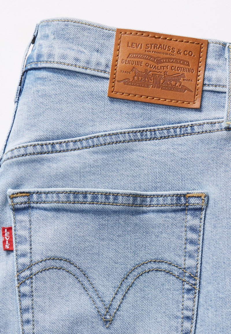Jeans en denim bleu clair avec un patch en cuir portant un texte en relief. Poches à double couture et petite étiquette rouge sur le côté.
