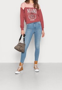 Sudadera color burdeos y rosa con la impresión "MIAMI 92 COLLEGE", combinada con vaqueros ajustados de color azul claro, zapatillas de caña alta color marrón claro y un bolso de cuadros.