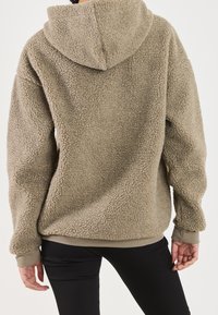 Fleece-hættetrøje i lys taupe med en tekstureret, blød overflade og en rund hætte. Har ribkantede manchetter og en afslappet pasform. Sorte bukser er synlige nedenunder.