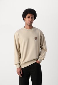 Felpa beige realizzata in tessuto morbido, con una grafica di un orso bruno e il testo "MAKIA" sul petto sinistro. Polsini a coste e scollatura a girocollo.