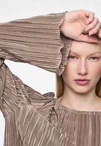 Haut plissé marron en tissu texturé, avec des poignets volantés et un col froncé. Le mannequin prend la pose avec un bras levé, dévoilant les détails des manches.