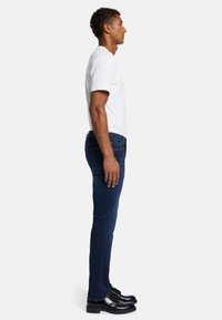Jeans blu scuro con vestibilità dritta, indossati con una t-shirt bianca a maniche corte e scarpe nere stringate. La vista laterale mostra una silhouette snella.