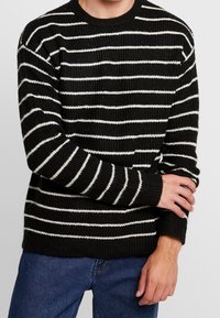 Czarnobiały sweter w paski, wykonany na drutach, z okrągłym dekoltem, długimi rękawami i ściągaczami na rękawach, noszony na dżinsach. Teksturowana tkanina.