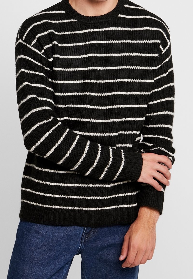 Czarnobiały sweter w paski, wykonany na drutach, z okrągłym dekoltem, długimi rękawami i ściągaczami na rękawach, noszony na dżinsach. Teksturowana tkanina.