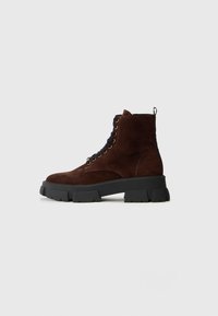 Stivale alla caviglia in suede marrone scuro con lacci, suola alta nera robusta, vista laterale su uno sfondo chiaro e semplice.