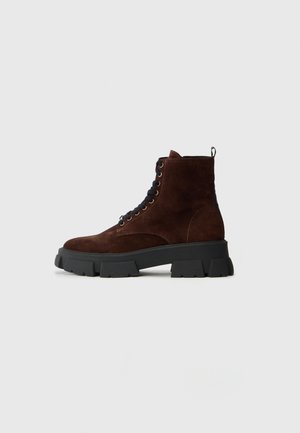 Stivale alla caviglia in suede marrone scuro con lacci, suola alta nera robusta, vista laterale su uno sfondo chiaro e semplice.