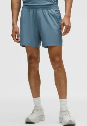 Man draagt lichtblauwe sportshorts, witte sneakersokken en witte hardloopschoenen, staand voor een effen achtergrond.