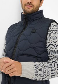 Marineblauer gesteppter Weste mit hohem Kragen und schwarzem Reißverschluss, getragen über einem gemusterten grauen Pullover mit geometrischen Designs in Marineblau.