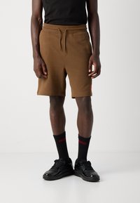 Bărbat îmbrăcat în pantaloni scurți maro cu șnur, maiou negru, șosete negre cu logo roșu și adidași negri, stând în fața unui fundal simplu.