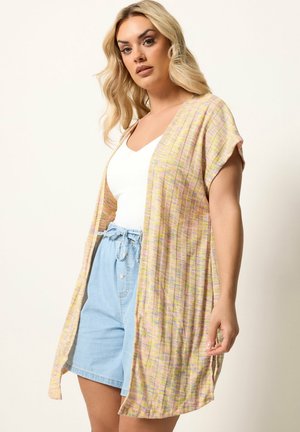 Lyshåret kvinde iført en hvid tanktop, lyseblå shorts med højtaljet pasform og en lang, åben cardigan med pastelgule og -lyserøde striber.