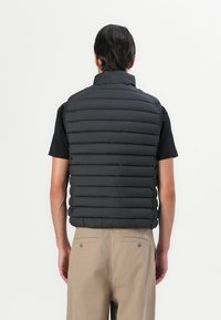Sort polstret vest med vandrette quiltede sektioner, høj krave og let materiale. Båret over en sort skjorte og lyse beige bukser.