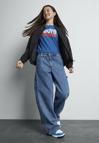 Losse blauwe denim jeans met een hoge taille, gecombineerd met een blauwe graphic t-shirt en een zwarte bomberjack, afgewerkt met wit-blauwe sneakers.