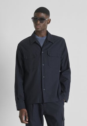 Man draagt een donkerblauwe jas met knopen en klepzakken, bijpassende broek, blauw overhemd en grote donkere zonnebril, staand tegen een grijze achtergrond.