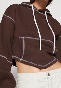 Brown cropped hoodie featuring white stitching details and a front pocket, with a drawstring hood and soft fabric texture.