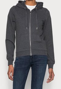 Grijze zip-up hoodie van zachte stof, voorzien van een trekkoordkap, voorkantzakken en geribde manchetten. Gecombineerd met donkere jeans.