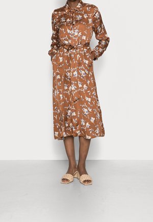 Robe chemise mi-longue fleurie couleur rouille, manches longues, poche sur la poitrine, ceinture nouée à la taille, portée avec des sandales à talon bloc tressé beige.