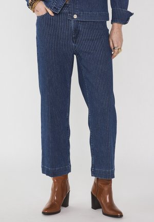 Jeans cropped a righe blu abbinati a stivaletti alla caviglia in pelle marrone con tacco a blocco, accessoriati con anelli e braccialetti dorati.