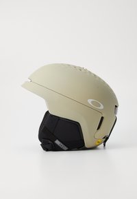 Casco de esquí de color beige claro con acabado mate, acolchado negro, agujeros de ventilación en la parte superior y correas ajustables. Presenta el logotipo de la marca en el lateral.