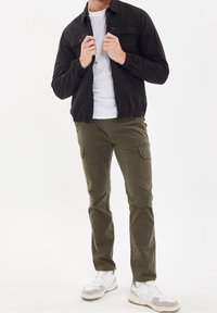 Veste noire à fermeture éclair avec poignets élastiques, T-shirt blanc, pantalon cargo vert olive avec poches, et baskets blanches avec accents gris.