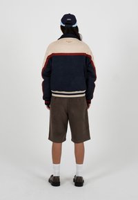 Personne portant une veste à blocs de couleurs, un short marron, des chaussettes blanches, des chaussures noires et une casquette de baseball bleu marine et bleu clair, debout, vue de dos.