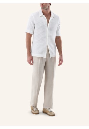 Mann trägt weißes kurzärmliges Strickhemd mit offenem Kragen, beige weite Hose und beige Schnürsneaker, steht vor hellem Hintergrund.