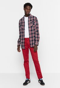Geruite shirt in navy, rood en wit met twee borstzakken; rode slim-fit broek; zwarte sneakers met witte accenten. Witte t-shirt eronder.