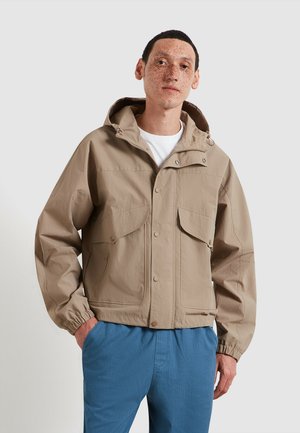 Giovane con capelli corti e scuri e lentiggini indossa una giacca leggera marrone chiaro con cappuccio sopra una camicia bianca e pantaloni blu, in piedi con le mani nelle tasche.