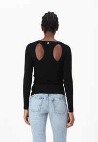 Svart ribbet langermet topp med åpen ryggdetalj, som har to utskårne seksjoner og en liten logo på øvre rygg. Kombinert med lyseblå jeans.