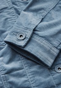 Détail d'une veste bleu clair avec une texture en velours côtelé, des manches à boutons et des coutures visibles. Les boutons comportent un logo embossé.