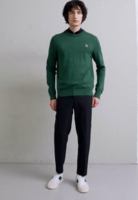 Green knitted sweater with a round neckline, black tapered trousers, and white sneakers with a navy accent. Positioned against a plain backdrop.