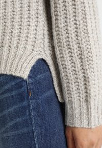 Jasnoszary sweter z dzianiny o prążkowanej fakturze i bocznych rozcięciach, zestawiony z ciemnoniebieskimi dżinsami. Zbliżenie na detale rękawa i pasa.