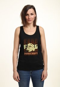 Canottiera nera con un design di Minecraft caratterizzato da elementi grafici beige e marroni, inclusi una scatola e testo pixelato. Visibili anche jeans di denim.