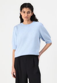Sweater azul claro com mangas curtas e bufantes, decote redondo e bordas caneladas. Usado sobre calças pretas, com um tecido macio e texturizado.