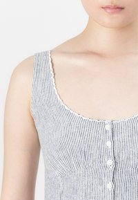 Close-up do torso superior de uma pessoa vestindo uma blusa de botões sem mangas, azul e branca com riscas e com um acabamento de gola em forma de escama.