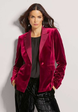 Bordeaux fluwelen blazer met een satijnen pieklabel, twee voorzakken en een enkele knoopsluiting; gedragen over een zwarte top en gestructureerde zwarte broek.