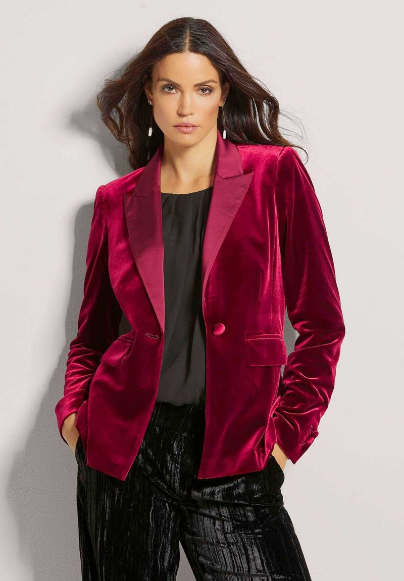 Bordeaux fluwelen blazer met een satijnen pieklabel, twee voorzakken en een enkele knoopsluiting; gedragen over een zwarte top en gestructureerde zwarte broek.