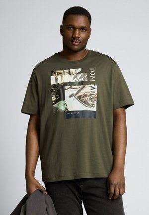 Man draagt een olijfgroen T-shirt met een afbeelding van een auto en tekst, houdt een donkere jas in zijn linkerhand, staand tegen een effen achtergrond.