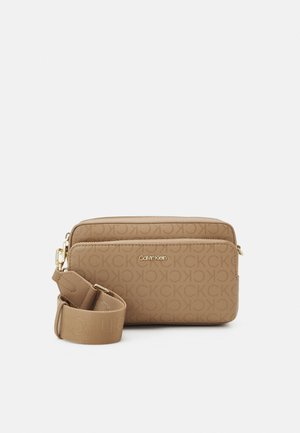 Beżowa torba na ramię Calvin Klein z monogramem CK, złote logo na przedniej kieszeni i szeroki regulowany pasek zwinięty obok niej.